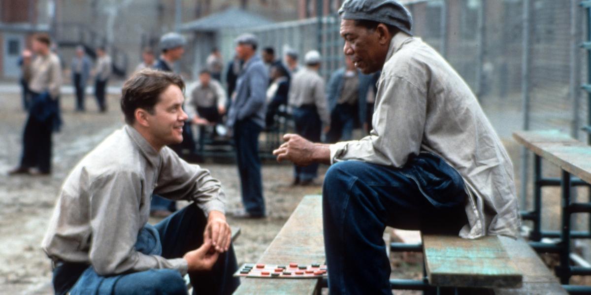 Tim Robbins and Morgan Freeman sit on a bench outside playing checkers and talking in a scene from the film